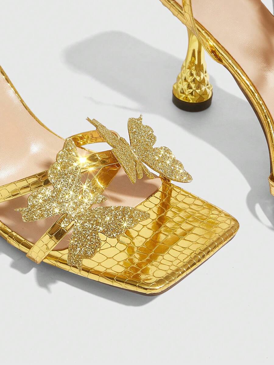 Glossy gold high heels featuring a sculptural heel, open-toe design, ankle strap buckle closure, and sparkling rhinestone butterfly embellishments.