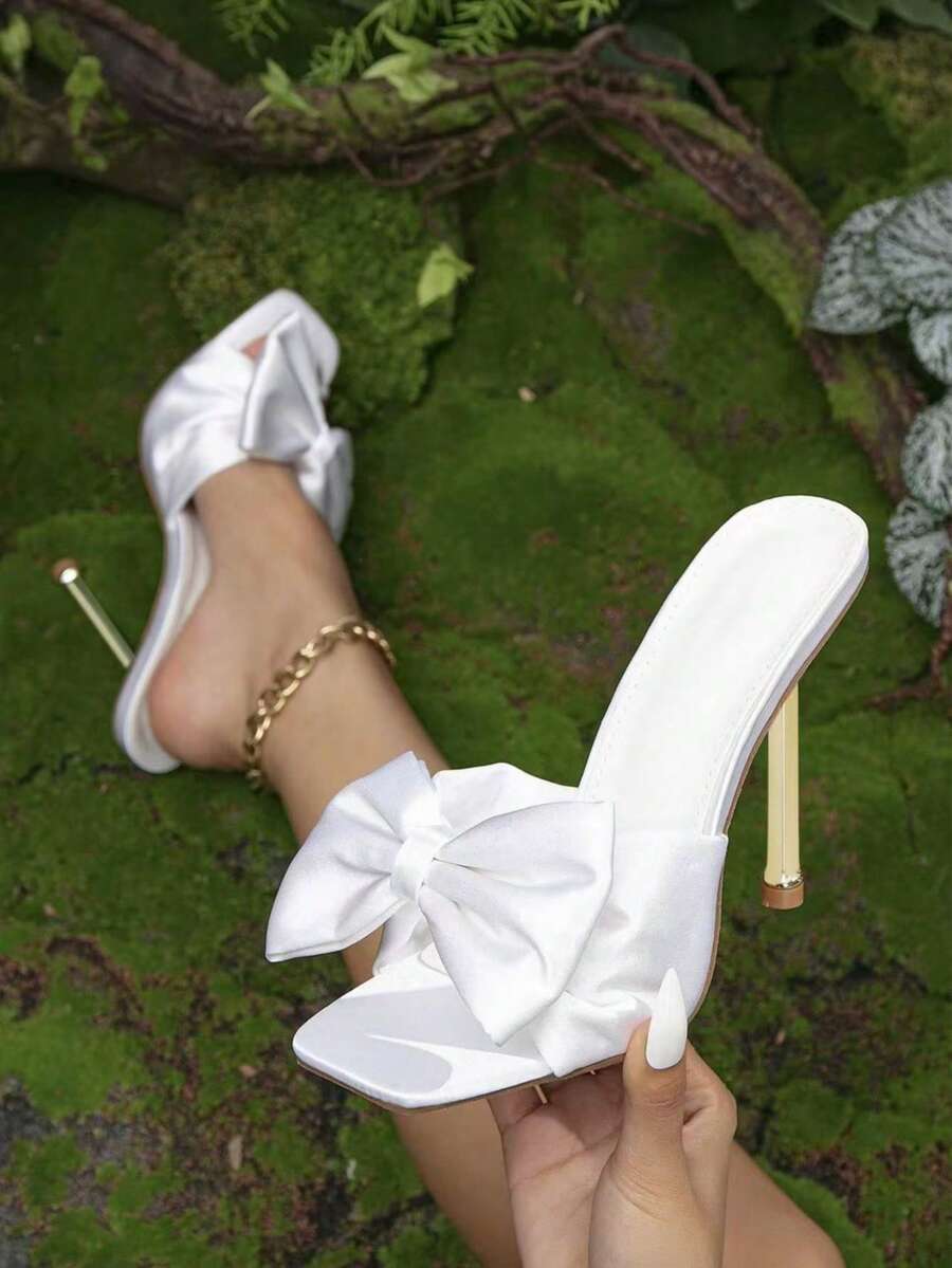 Women's black satin mule heels featuring a delicate bow detail, pointy open toe, and metallic electroplated stiletto heel.