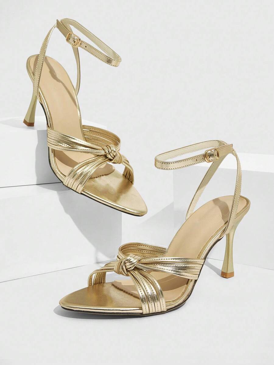 Open toe high heel sandals in champagne gold featuring a twist knot front strap, sleek stiletto heel, and adjustable ankle buckle strap.