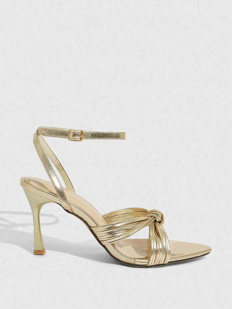 Open toe high heel sandals in champagne gold featuring a twist knot front strap, sleek stiletto heel, and adjustable ankle buckle strap.