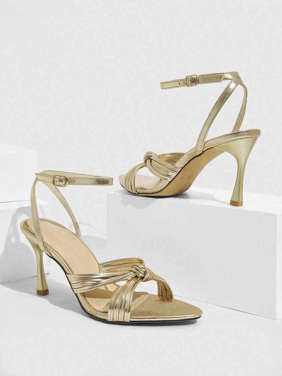 Open toe high heel sandals in champagne gold featuring a twist knot front strap, sleek stiletto heel, and adjustable ankle buckle strap.