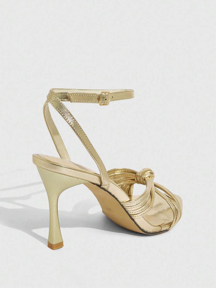 Open toe high heel sandals in champagne gold featuring a twist knot front strap, sleek stiletto heel, and adjustable ankle buckle strap.