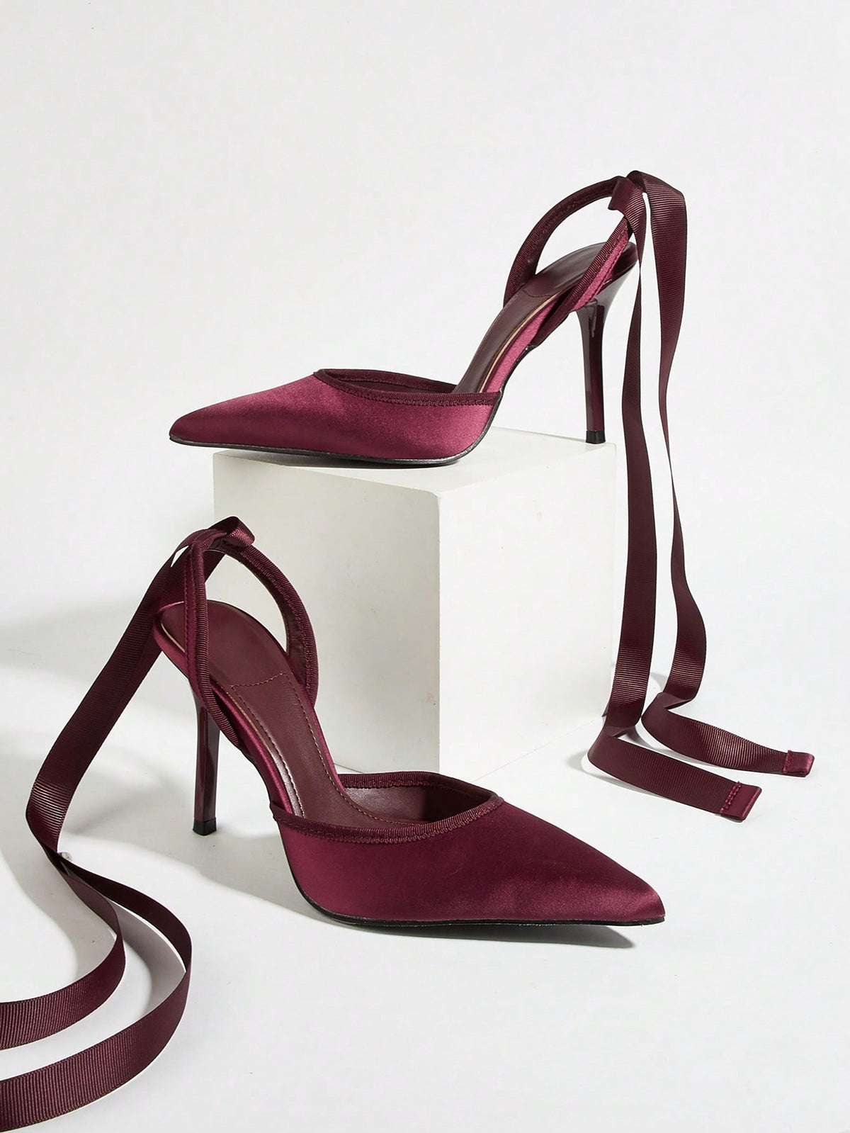 Burgundy satin pointed toe stiletto high heel pumps with a backless design and delicate bow accent.