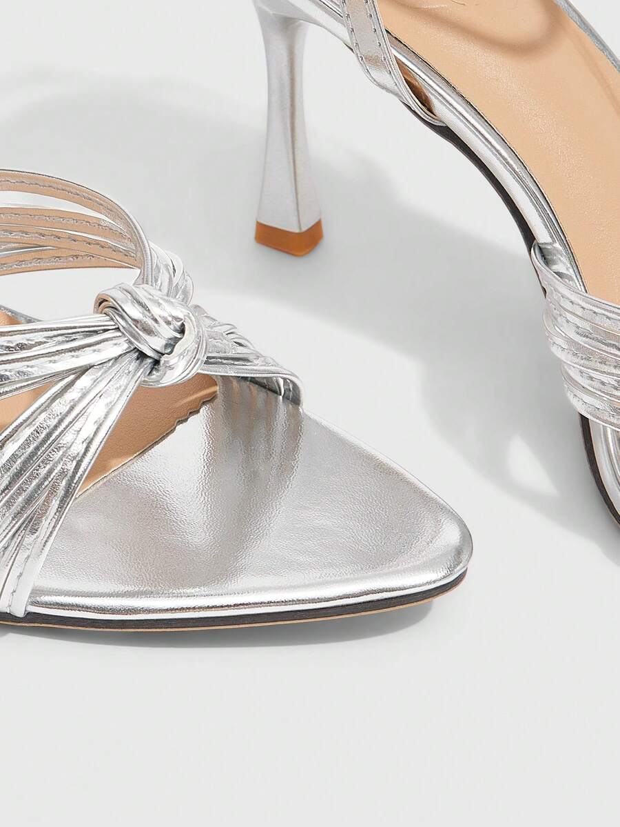 Open toe high heel sandals in champagne gold featuring a twist knot front strap, sleek stiletto heel, and adjustable ankle buckle strap.