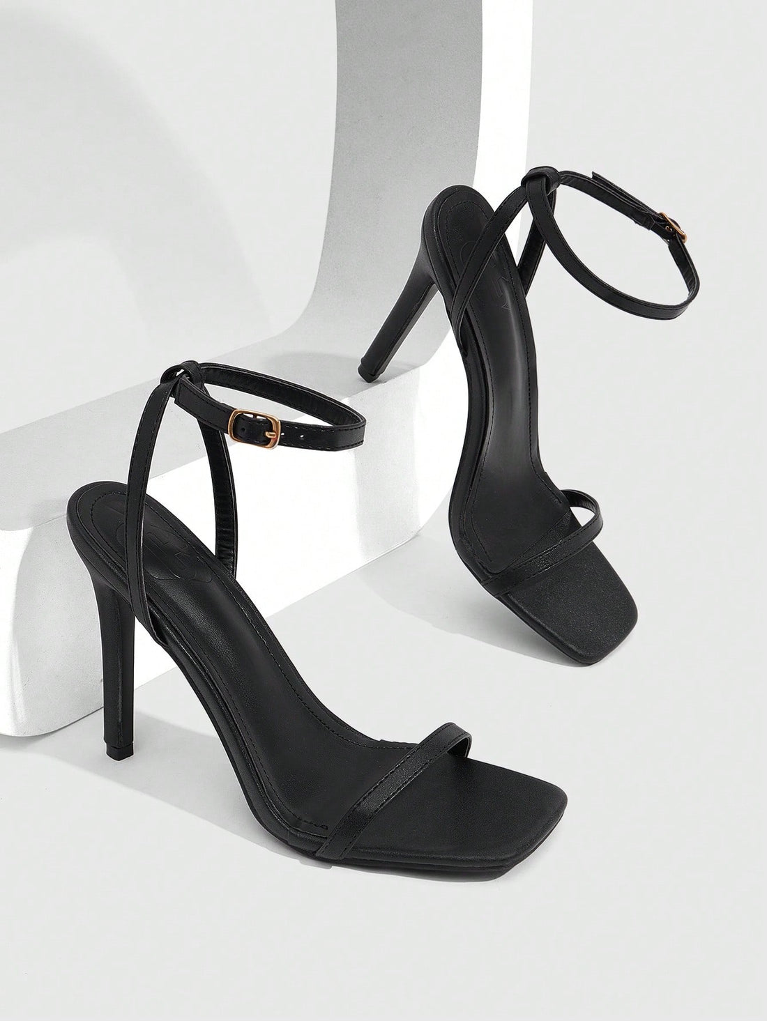 Women's minimalist black heeled sandals featuring an open toe, stiletto high heel, and adjustable ankle strap buckle closure.