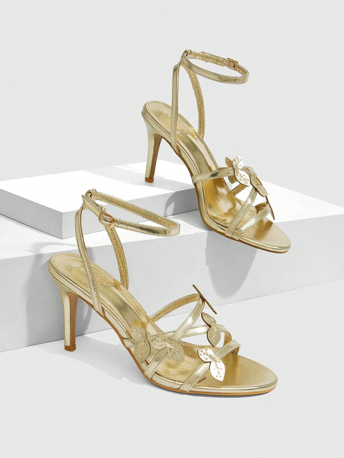 Metallic gold stiletto high heel sandals for women with criss-cross straps, leaf decor accents, and buckle ankle closure.