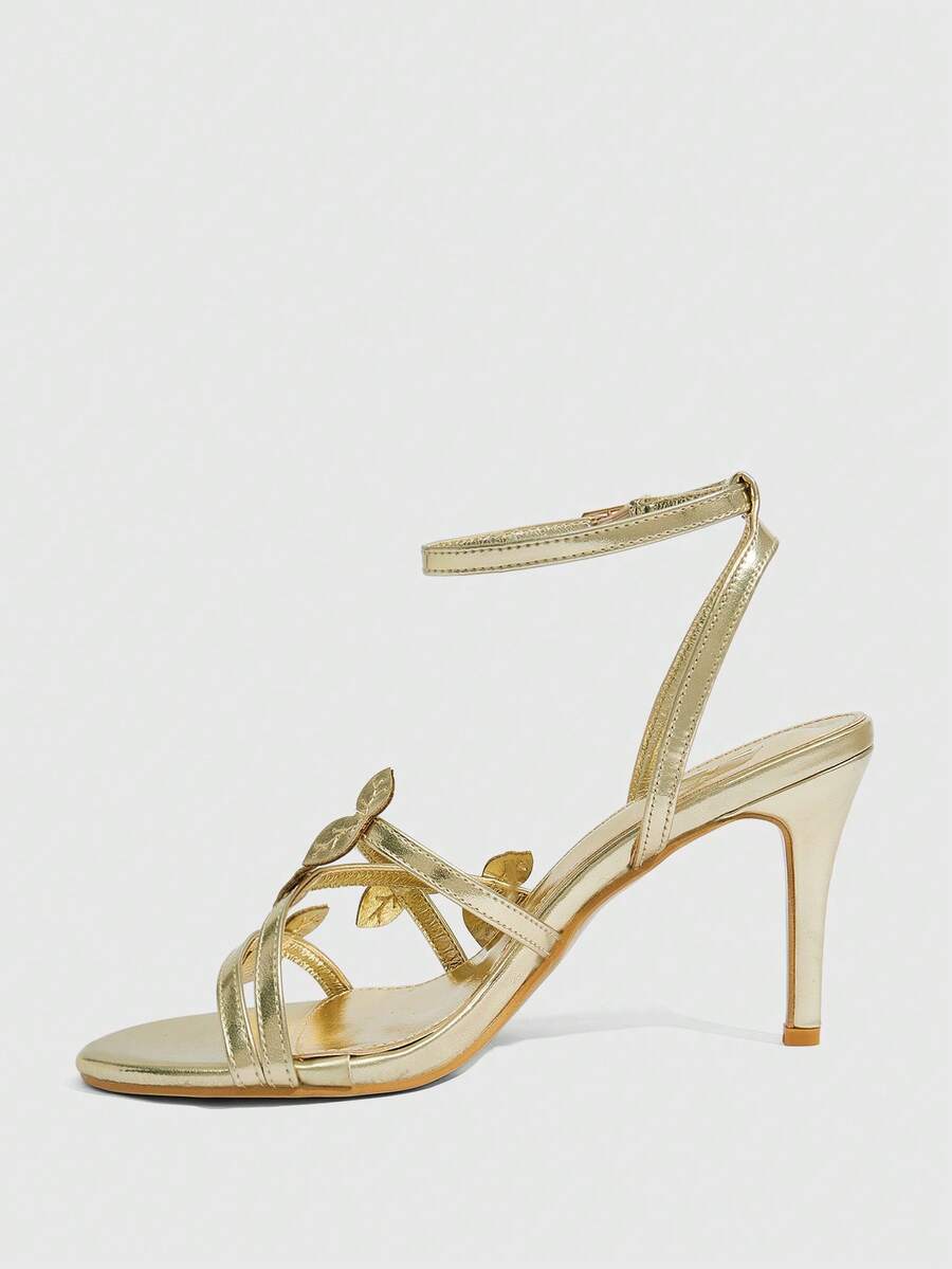 Metallic gold stiletto high heel sandals for women with criss-cross straps, leaf decor accents, and buckle ankle closure.