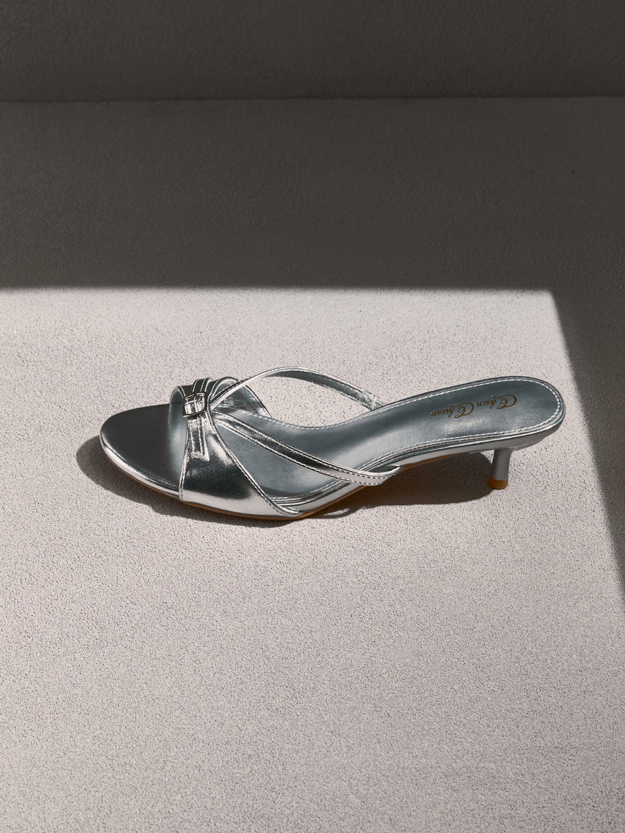 Pair of women's silver kitten heel sandals featuring criss-cross slip-on straps, an open round toe, and an elegant bow detail.