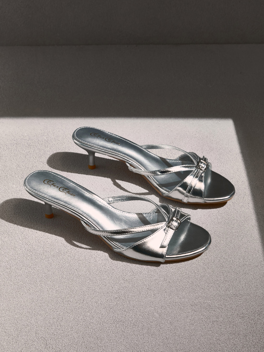 Pair of women's silver kitten heel sandals featuring criss-cross slip-on straps, an open round toe, and an elegant bow detail.