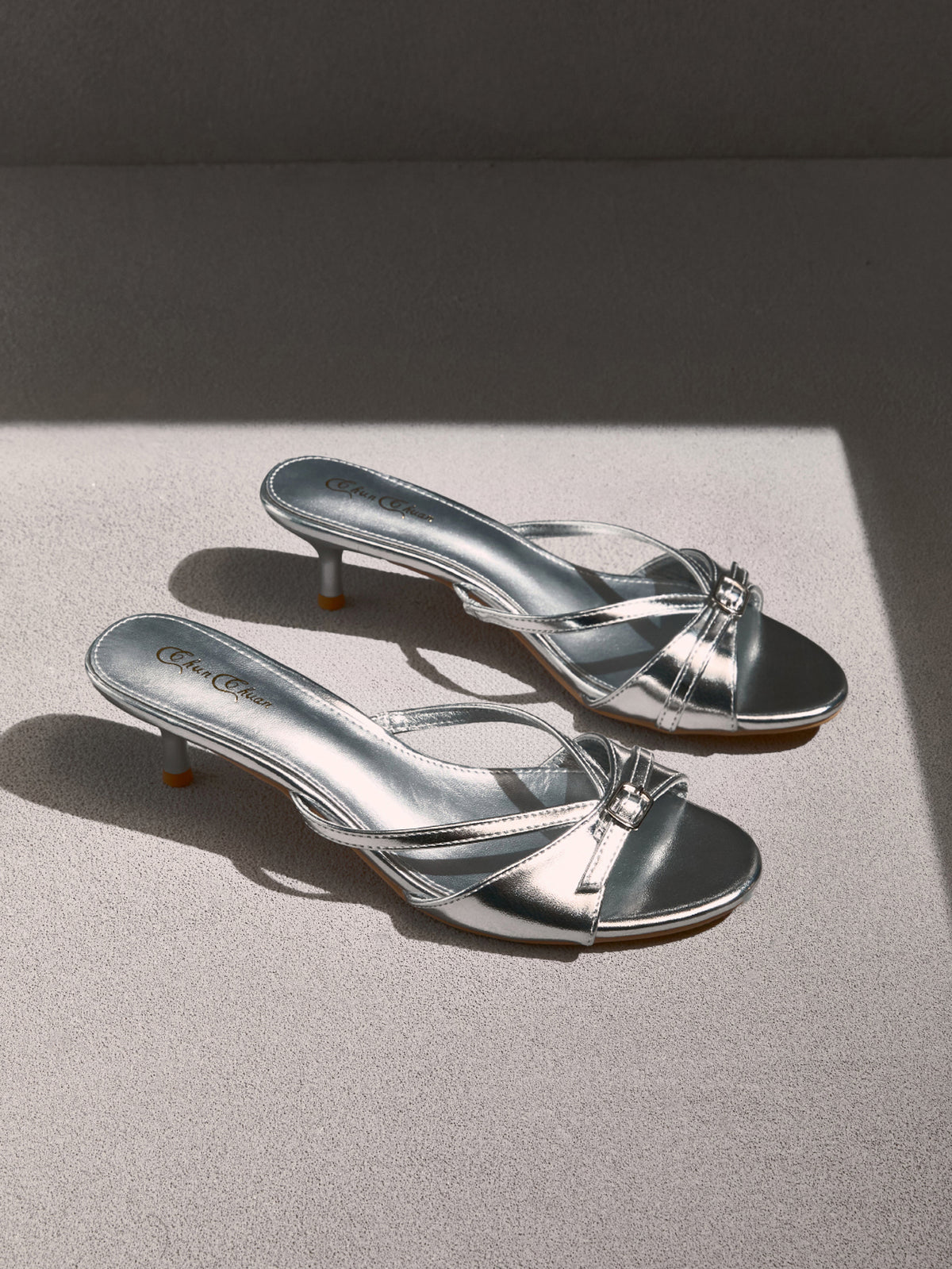 Pair of women's silver kitten heel sandals featuring criss-cross slip-on straps, an open round toe, and an elegant bow detail.