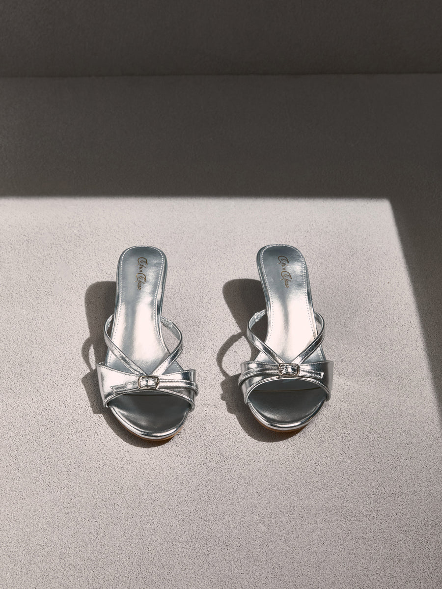 Pair of women's silver kitten heel sandals featuring criss-cross slip-on straps, an open round toe, and an elegant bow detail.
