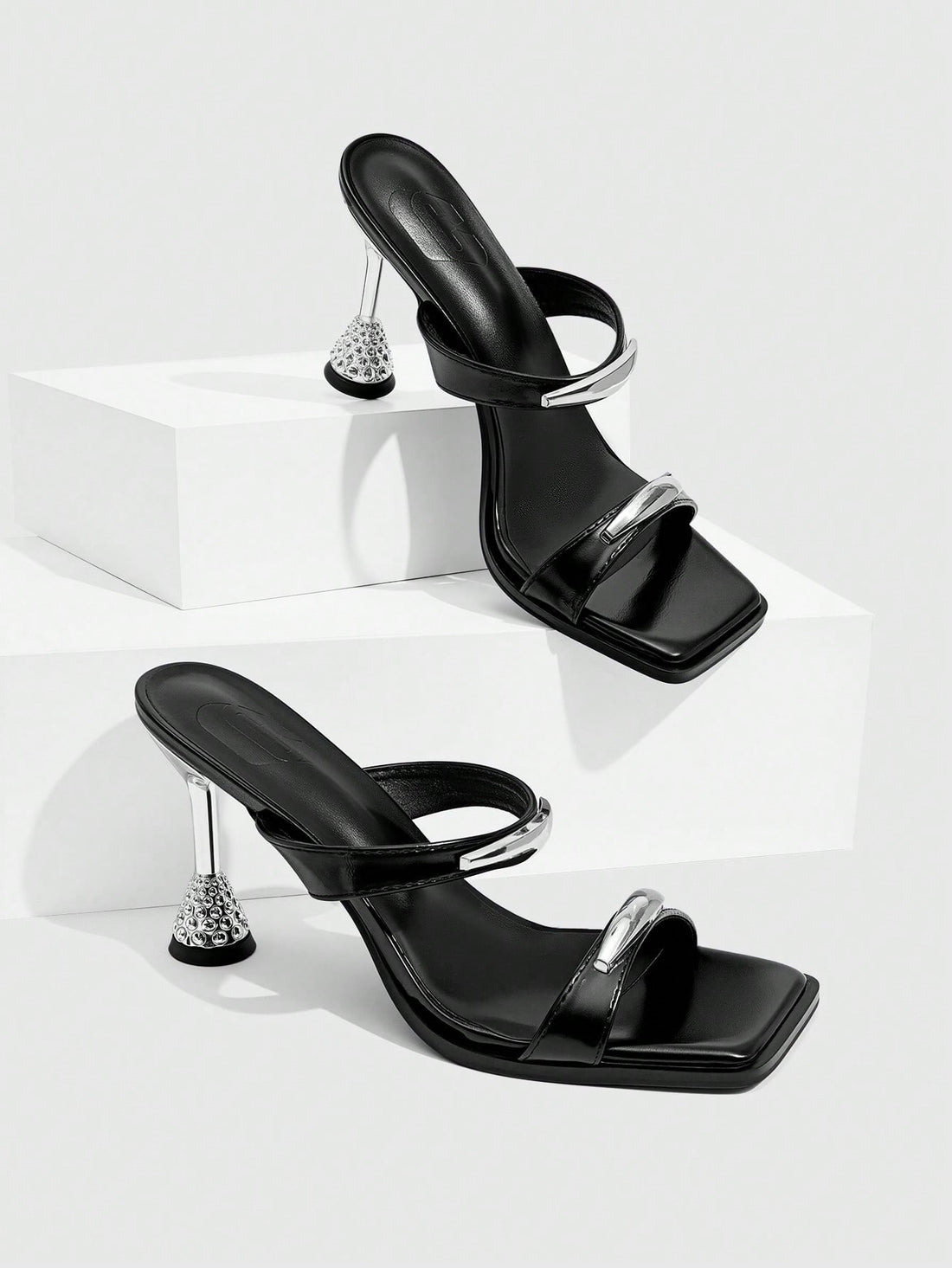 Women's black high-heeled sandals featuring an open square toe, stiletto heel, and decorative metal buckle strap detail.