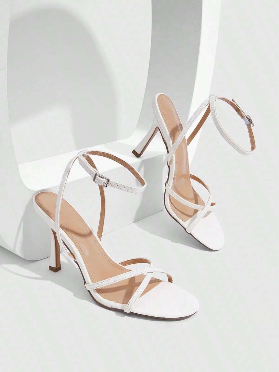 Pair of cream open-toe heeled sandals featuring crisscross front straps, a sleek stiletto high heel, and an adjustable ankle strap with buckle closure.