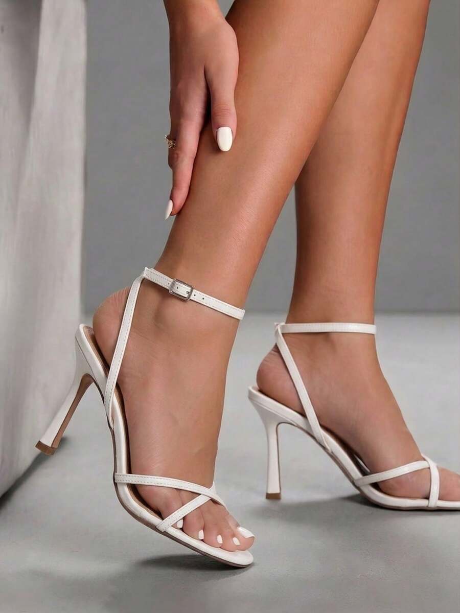 Pair of cream open-toe heeled sandals featuring crisscross front straps, a sleek stiletto high heel, and an adjustable ankle strap with buckle closure.