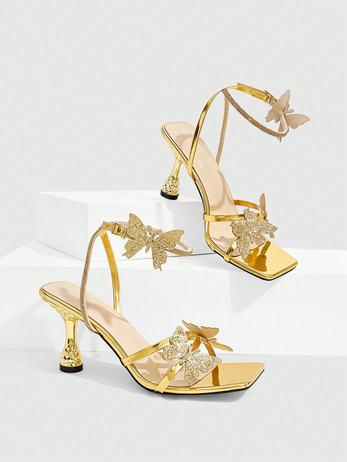 Glossy gold high heels featuring a sculptural heel, open-toe design, ankle strap buckle closure, and sparkling rhinestone butterfly embellishments.