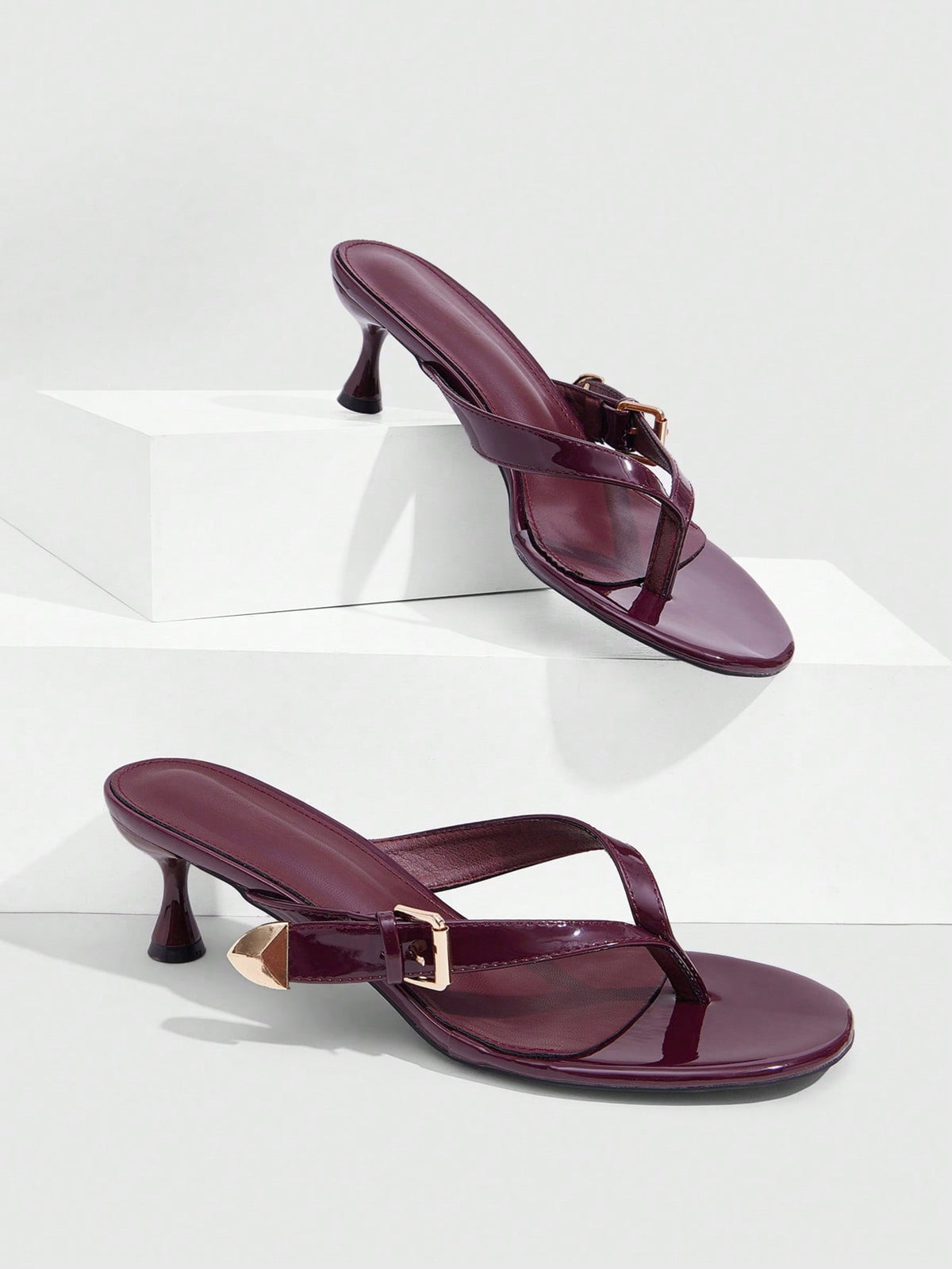 Women's patent leather heeled sandals featuring a toe post strap with metal buckle and mid pyramid heel.