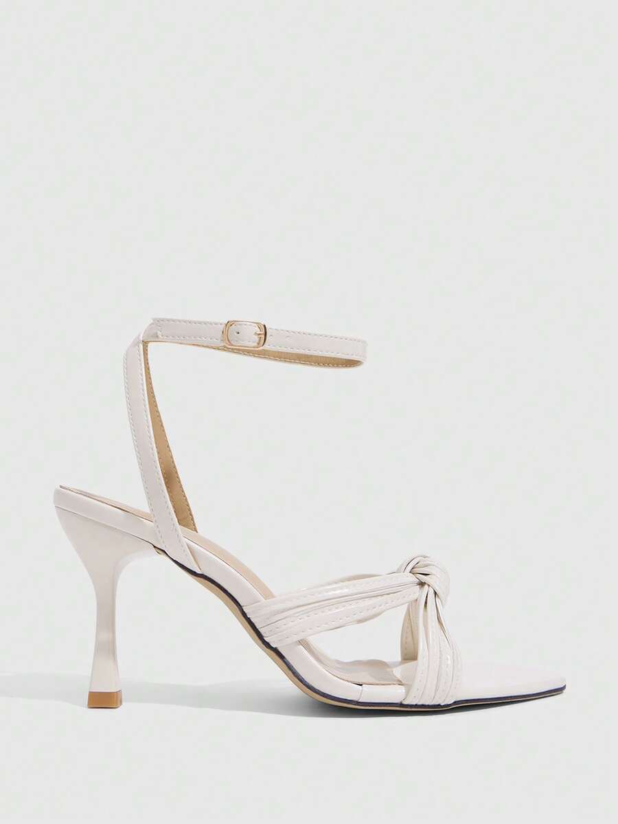 Open toe high heel sandals in champagne gold featuring a twist knot front strap, sleek stiletto heel, and adjustable ankle buckle strap.