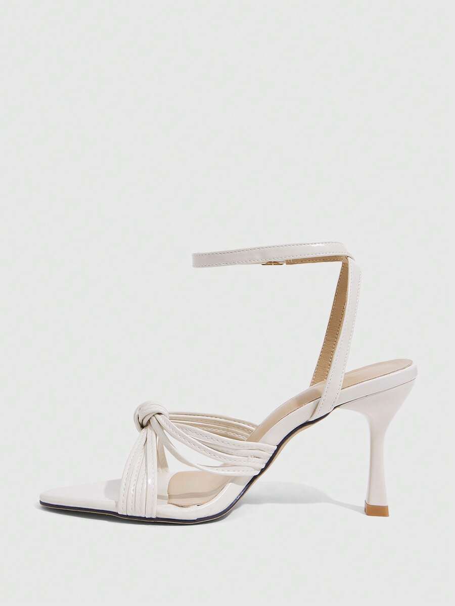 Open toe high heel sandals in champagne gold featuring a twist knot front strap, sleek stiletto heel, and adjustable ankle buckle strap.