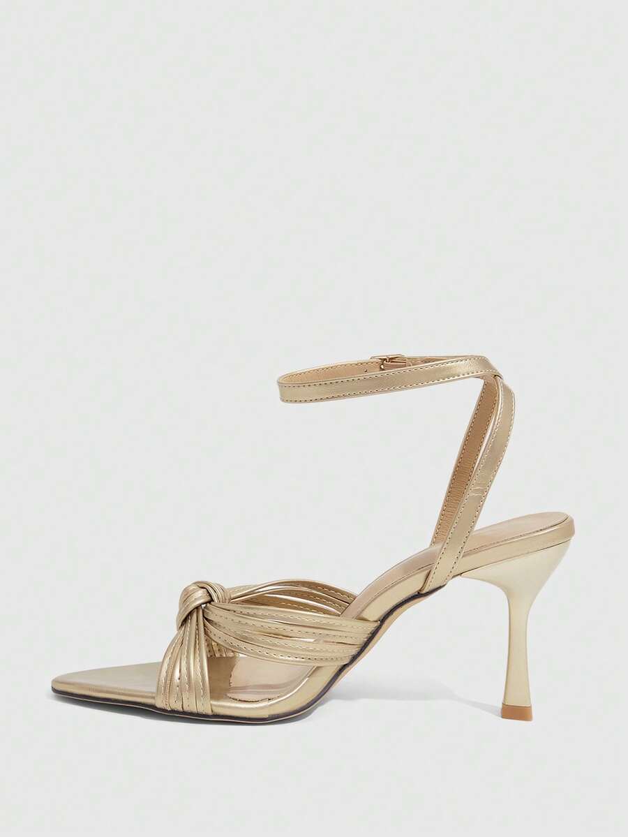 Open toe high heel sandals in champagne gold featuring a twist knot front strap, sleek stiletto heel, and adjustable ankle buckle strap.