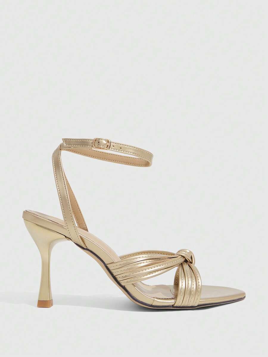 Open toe high heel sandals in champagne gold featuring a twist knot front strap, sleek stiletto heel, and adjustable ankle buckle strap.