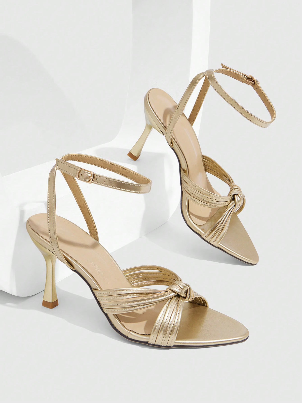 Open toe high heel sandals in champagne gold featuring a twist knot front strap, sleek stiletto heel, and adjustable ankle buckle strap.